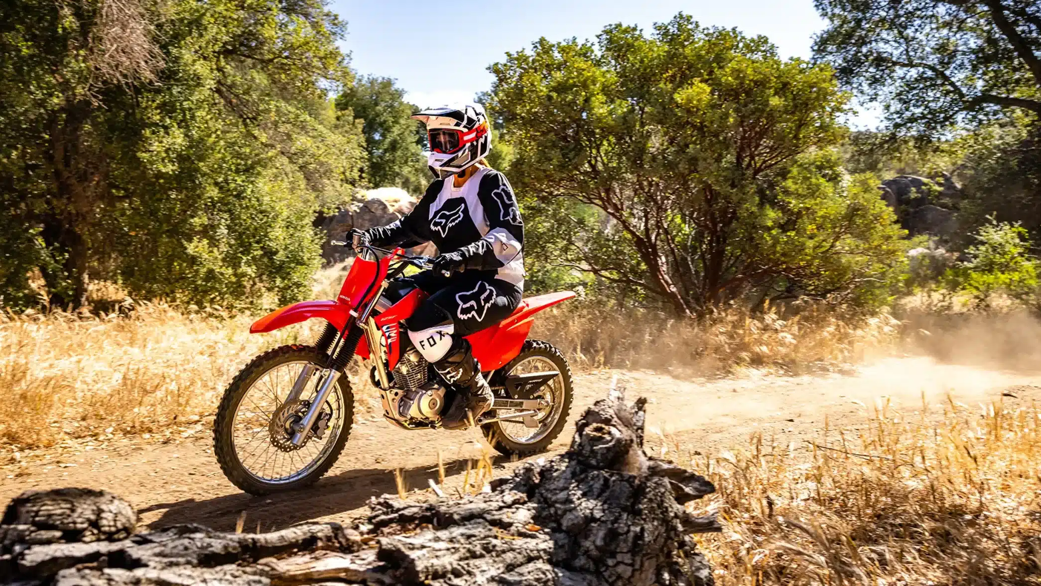 Honda CRF125F mit Fahrerin beim Offroad-Training auf staubigem Pfad, umgeben von Bäumen.
