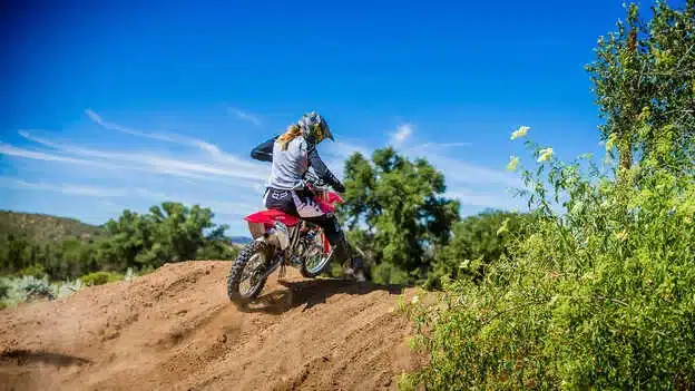 Motocross-Fahrer auf einer Honda CRF150R fährt eine sandige Piste mit üppiger Vegetation im Hintergrund.