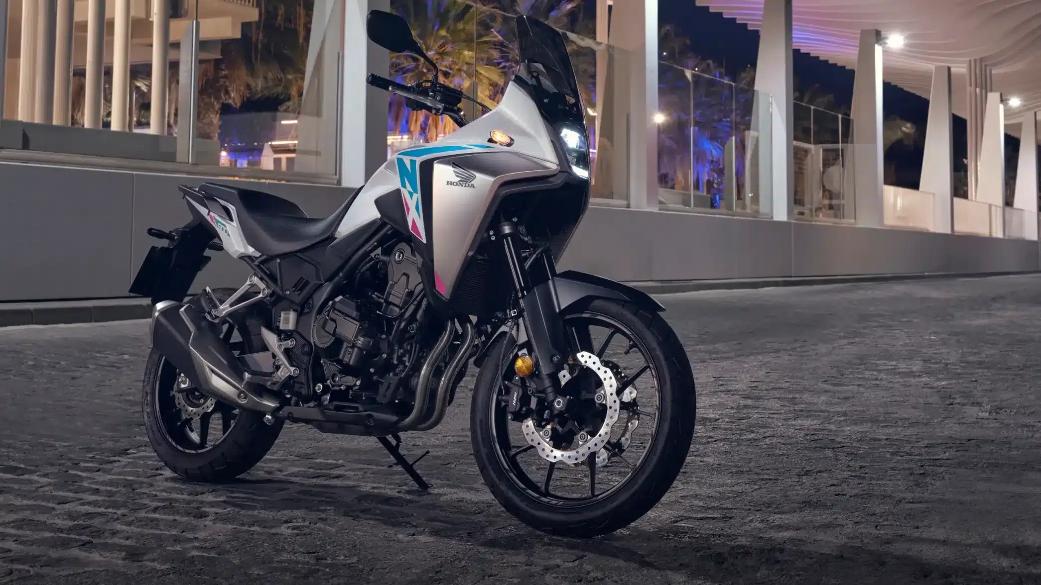 Honda NX500 Adventure-Motorrad auf Kopfsteinpflaster bei Nacht, seitliche Frontansicht, moderne Architektur im Hintergrund.