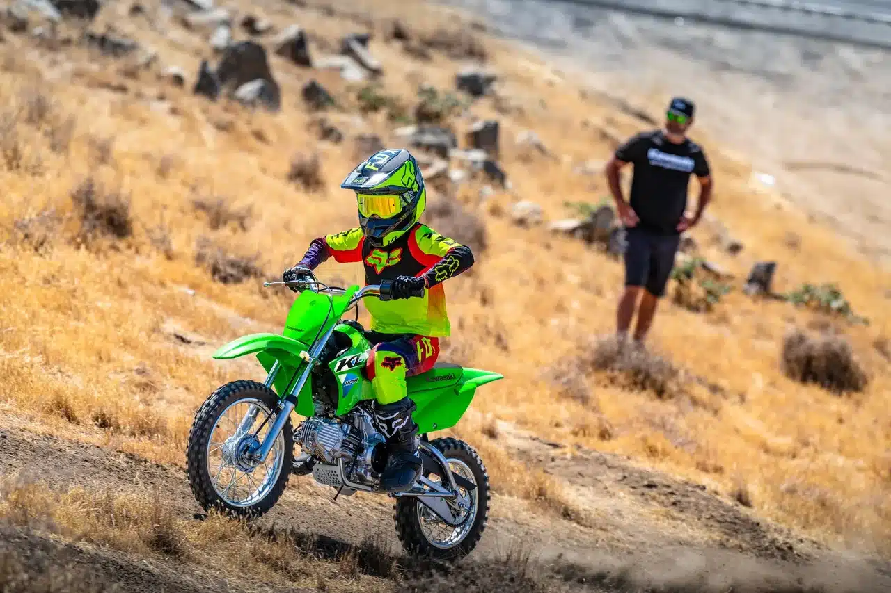 Junger Fahrer auf einer Kawasaki KLX110R 2026 im Motocross-Outfit fährt über eine trockene, felsige Landschaft.