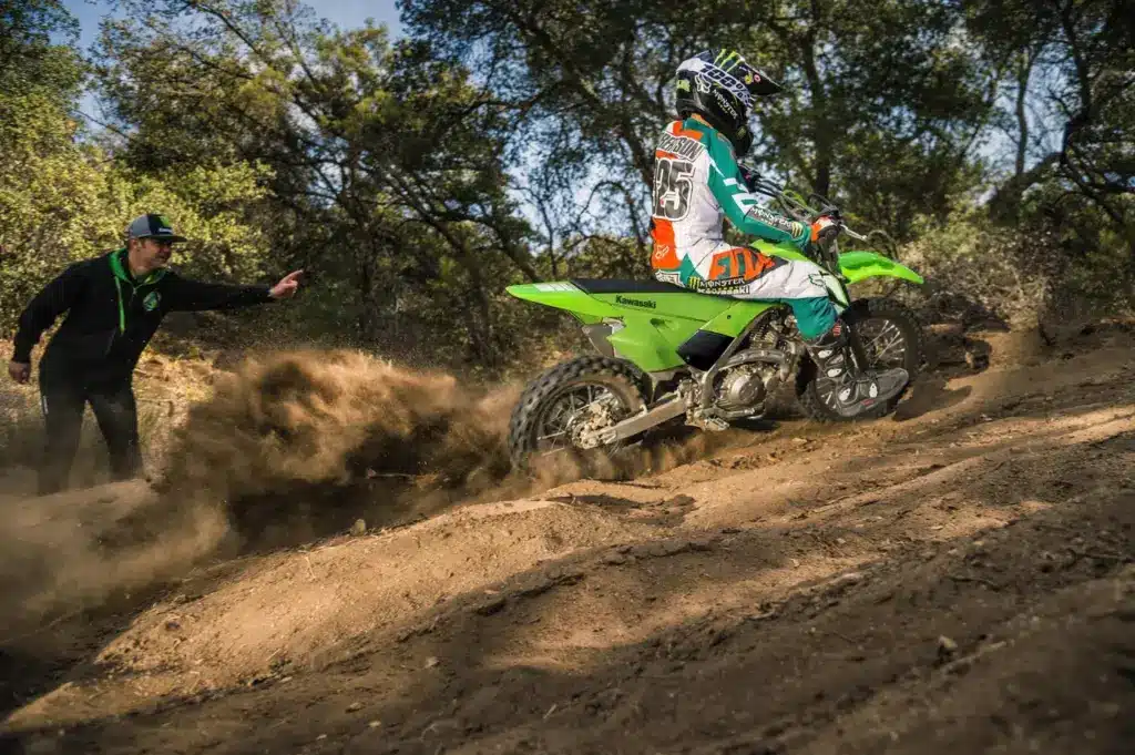 Fahrer auf einer grünen Kawasaki KLX140R fährt über eine staubige Offroad-Strecke, unterstützt von einer Person am Streckenrand