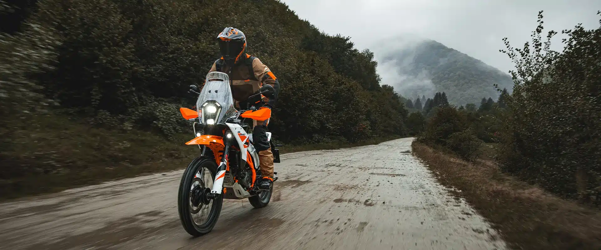 KTM 390 Adventure R 2026 Offroad-Motorrad fährt auf einer matschigen Schotterstraße mit bewaldeten Bergen im Hintergrund.
