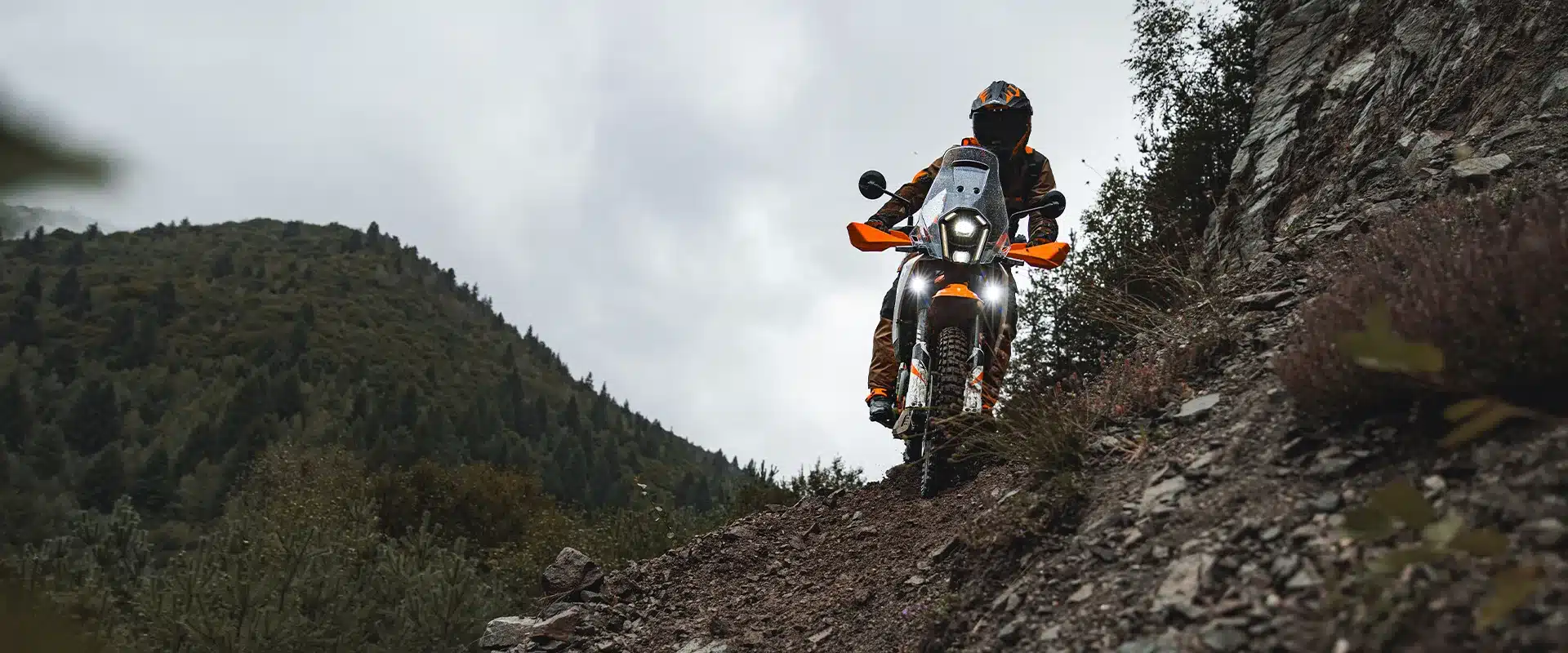 KTM 390 Adventure X 2026 fährt Offroad eine steile Geröllpiste hinunter, umgeben von Bergen und bewaldeten Hügeln.