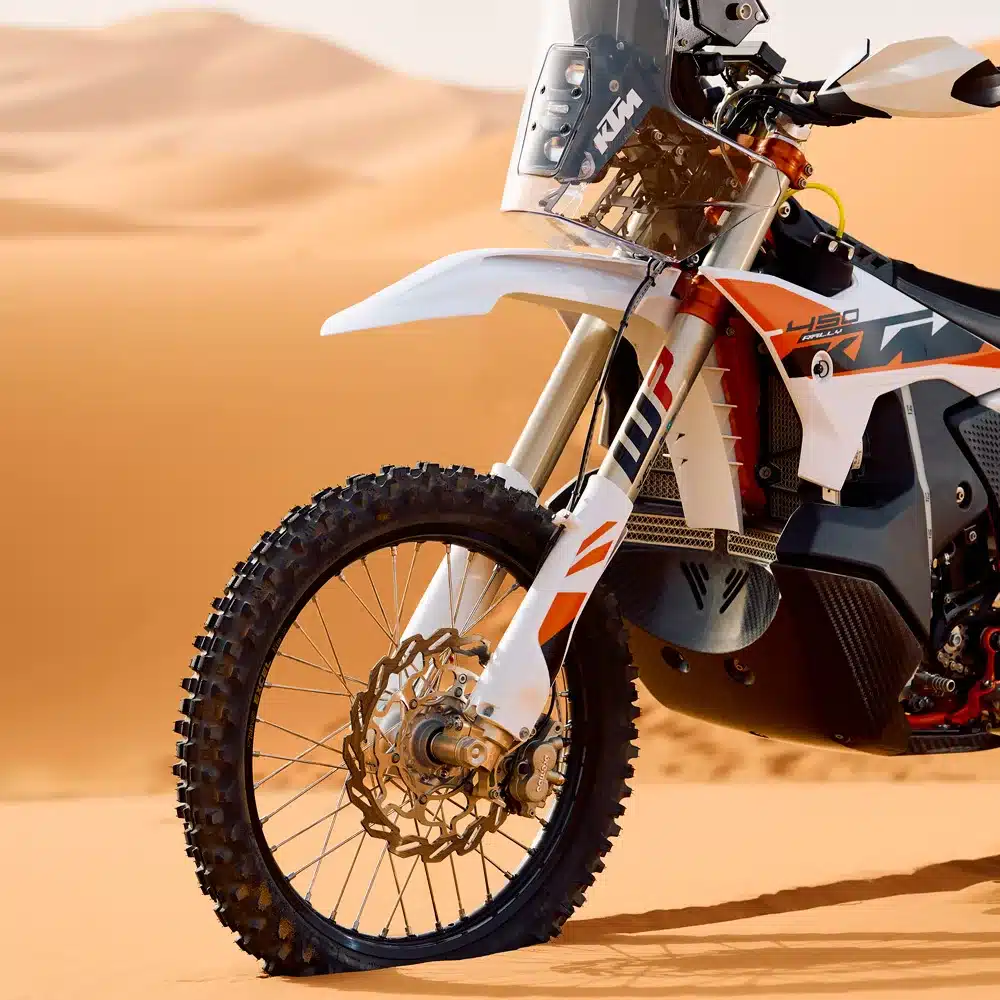 Nahaufnahme des Vorderrads und Kühlsystems der KTM 450 Rally Replica 2026 in einer Sandlandschaft.