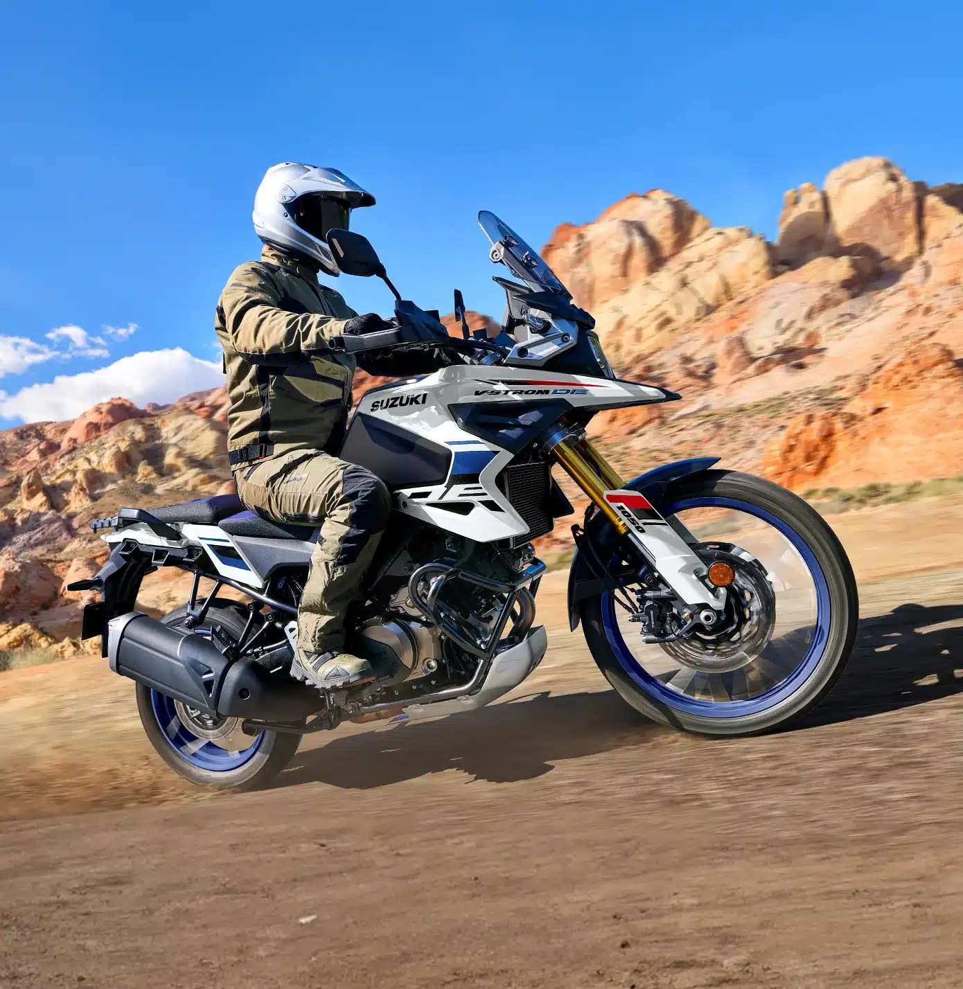 Abenteuer-Motorrad Suzuki V-Strom 1050DE fährt auf Schotterweg mit Fahrer in Offroad-Bekleidung vor Felsen.