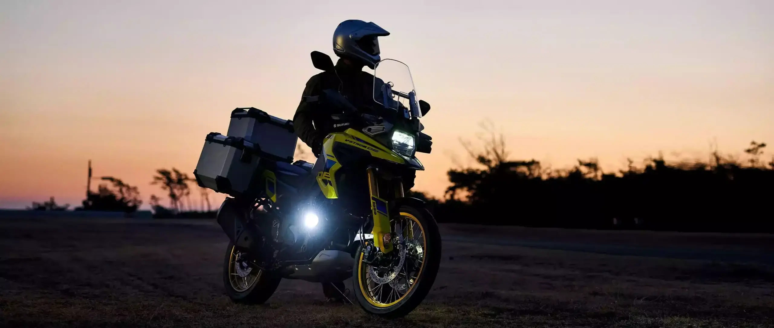 Gelbe Suzuki V-Strom 1050DE Travel Edition mit Aluminium-Koffern im Offroad-Einsatz bei Sonnenuntergang