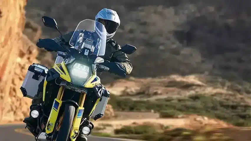 Suzuki V-Strom 1050DE Travel Edition in Gelb-Silber mit Aluminium-Koffern, gefahren von einem Motorradfahrer auf kurvenreicher Straße im Gebirge.