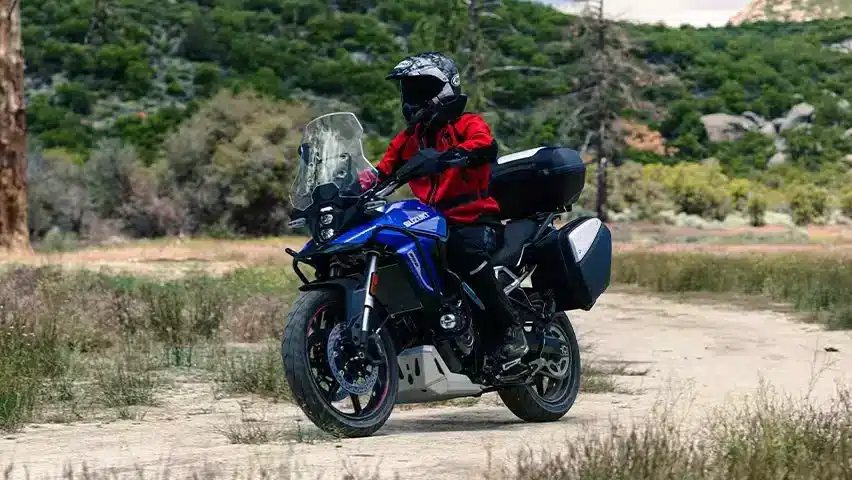 Blaues Suzuki V-Strom 800 Power Edition Adventure-Bike mit Gepäckkoffern auf einem Schotterweg, Fahrer in Motorradkleidung.