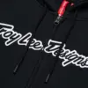 Nahaufnahme des schwarzen Troy Lee Designs Zip Up Hoodie Signature mit Reißverschluss und weißem Logo-Schriftzug.
