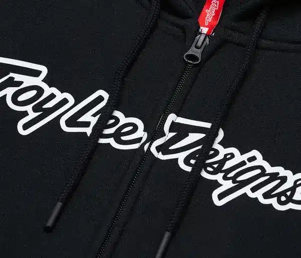 Nahaufnahme des schwarzen Troy Lee Designs Zip Up Hoodie Signature mit Reißverschluss und weißem Logo-Schriftzug.