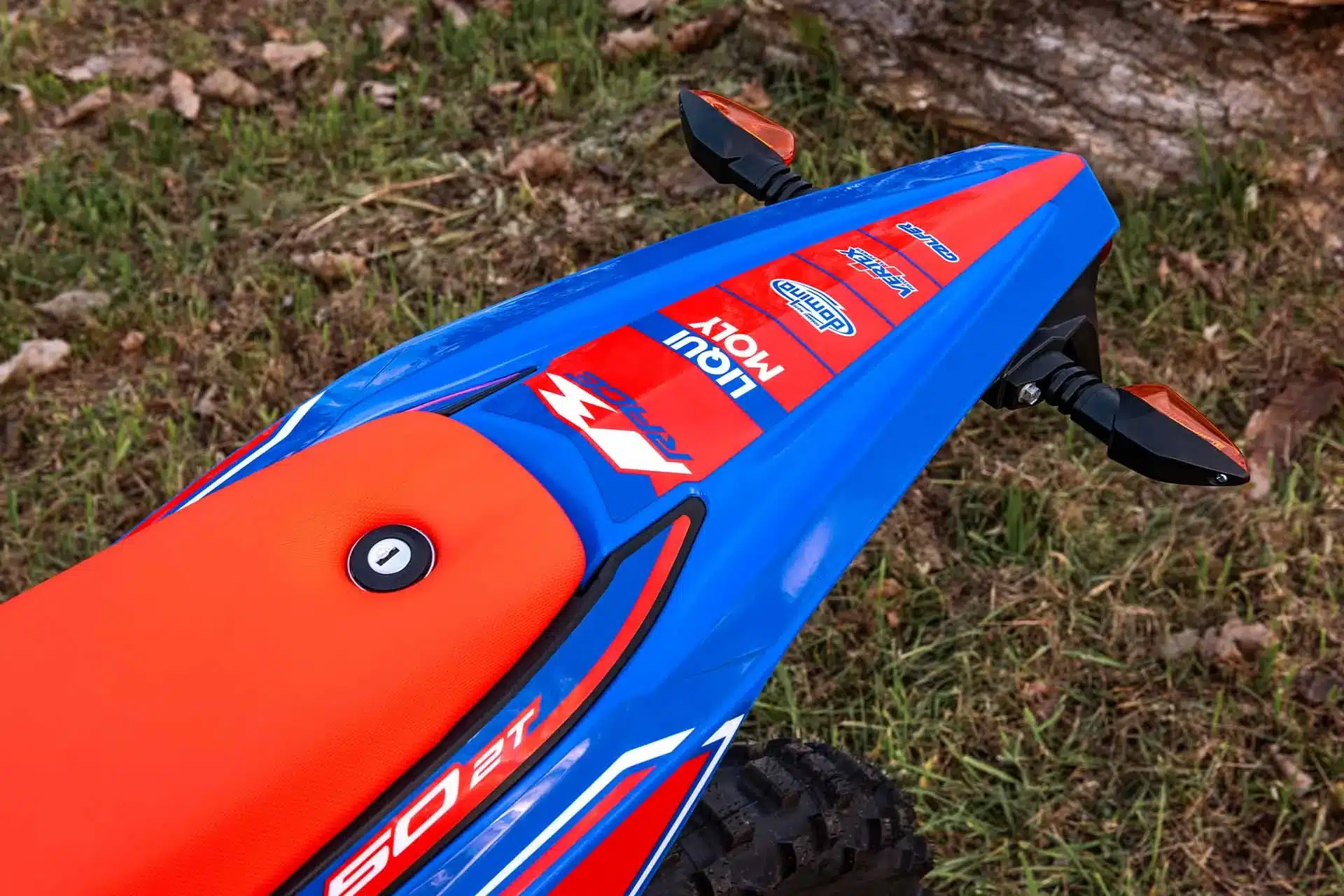Detailaufnahme des Hecks und der Sitzbank einer Beta RR 50 Race 2025 Enduro, blau-rot mit Sponsorenaufklebern auf Grasuntergrund.