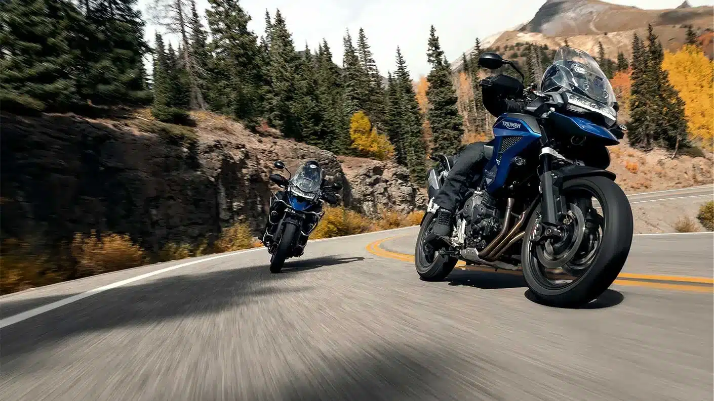 Triumph Tiger 1200 GT Explorer Motorrad fährt auf einer kurvigen Landstraße durch eine herbstliche Berglandschaft.