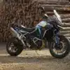 Triumph Tiger 900 Alpine Edition Motorrad mit Akrapovič-Schalldämpfer auf Schotterweg vor gestapelten Holzstämmen