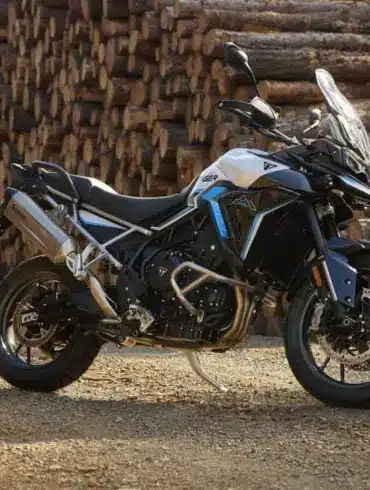 Triumph Tiger 900 Alpine Edition Motorrad mit Akrapovič-Schalldämpfer auf Schotterweg vor gestapelten Holzstämmen