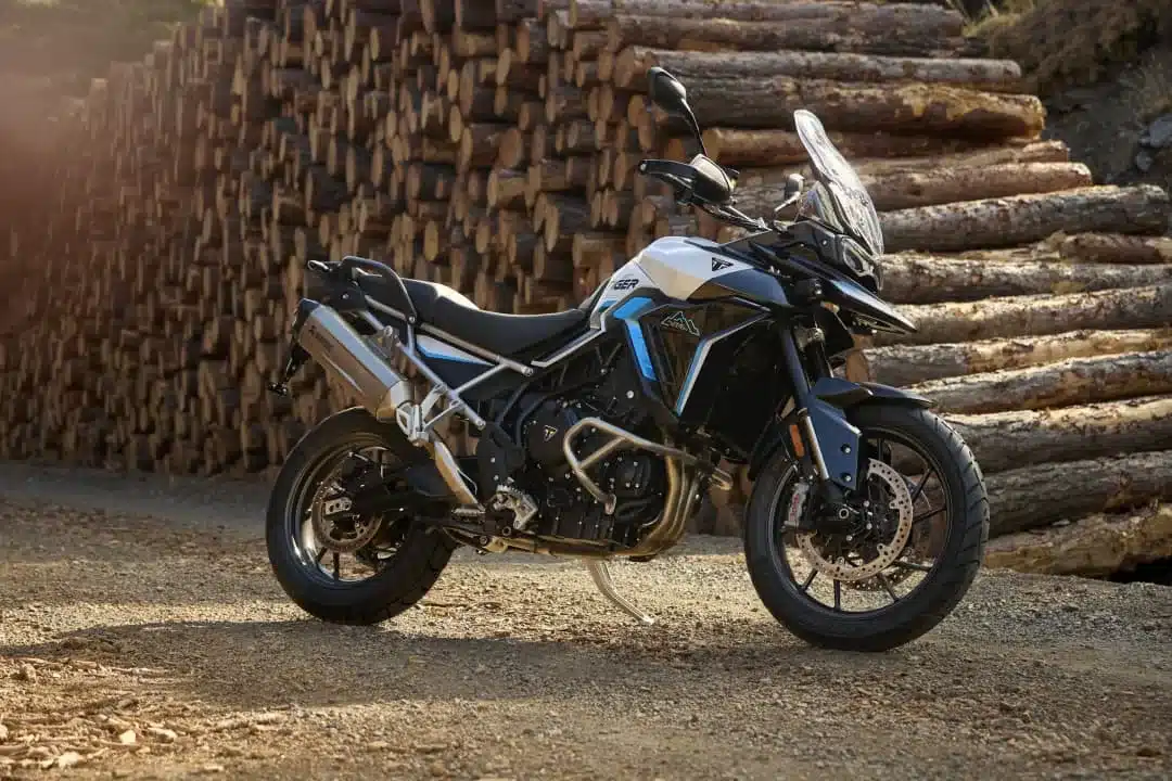 Triumph Tiger 900 Alpine Edition Motorrad mit Akrapovič-Schalldämpfer auf Schotterweg vor gestapelten Holzstämmen