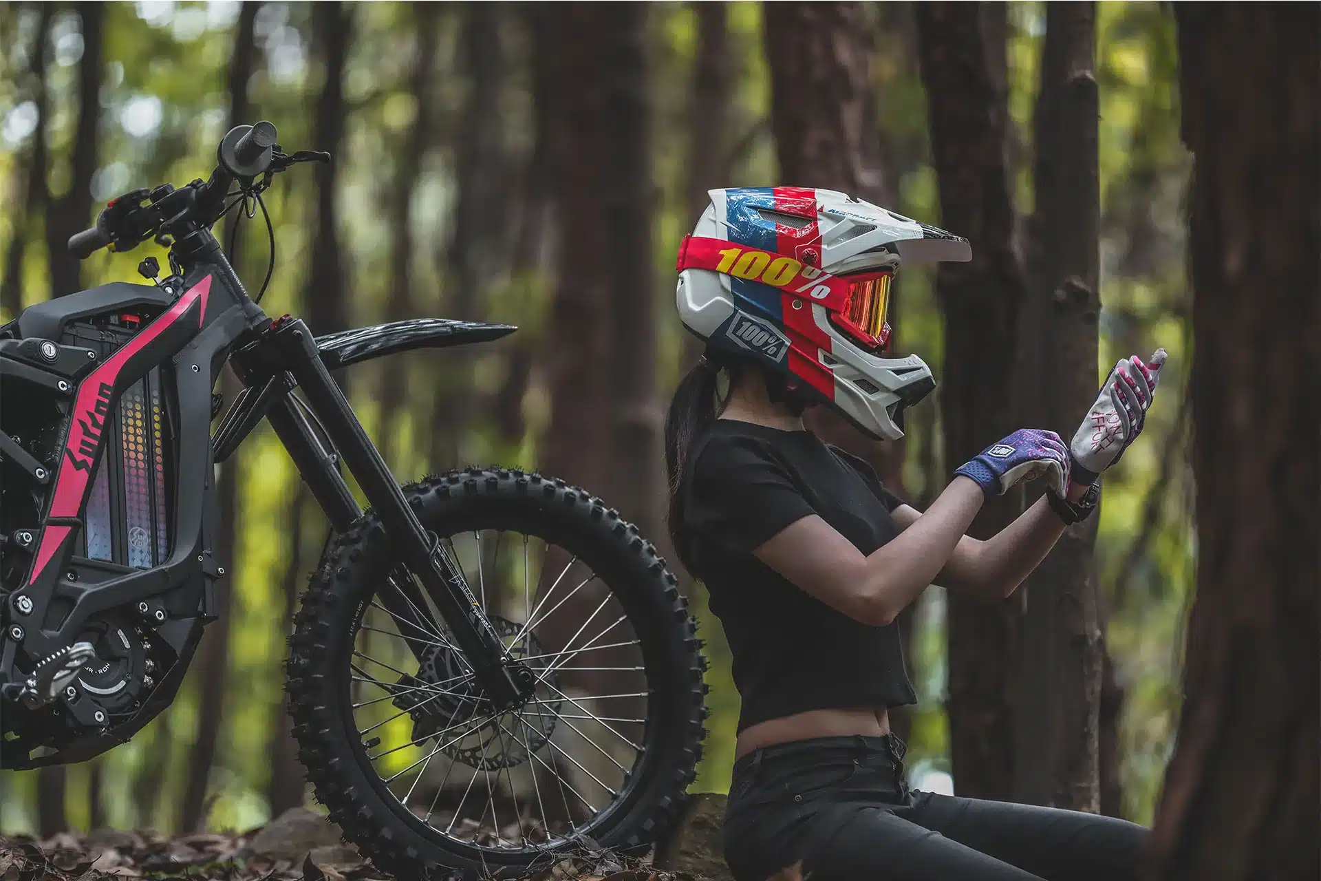 Fahrerin mit Helm sitzt im Wald neben einer Sur-Ron Light Bee S Elektro-Offroad-Motorrad und zieht Handschuhe an