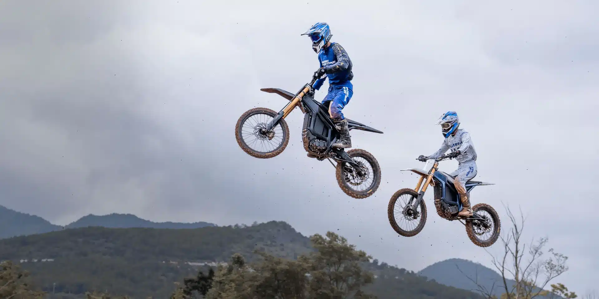 Zwei Talaria Komodo MX6 Elektro-Motorräder im Sprung auf Offroad-Strecke vor bewölktem Himmel.