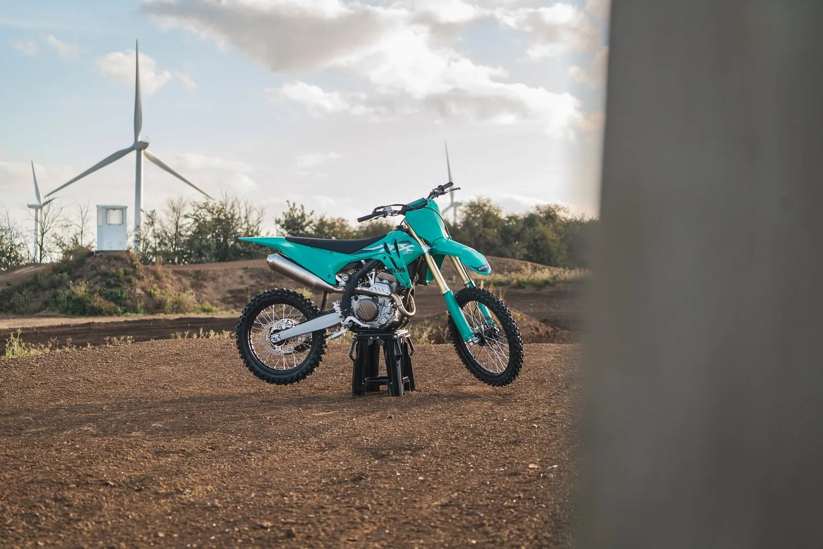 Türkise Kove MX250 Motocross-Maschine steht auf einer Offroad-Strecke im Freien, im Hintergrund Windräder.