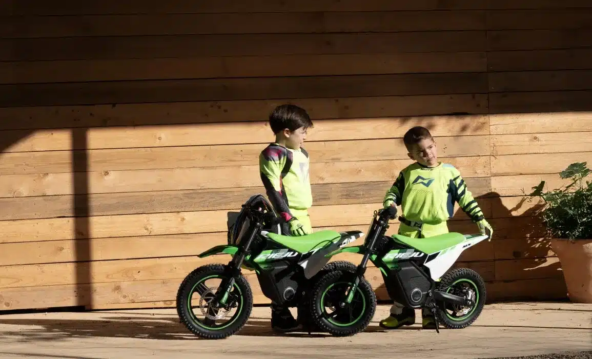 Zwei Kinder stehen in Motocross-Bekleidung neben grünen Rieju MX12 250W Elektromotorrädern vor einer Holzfassade.