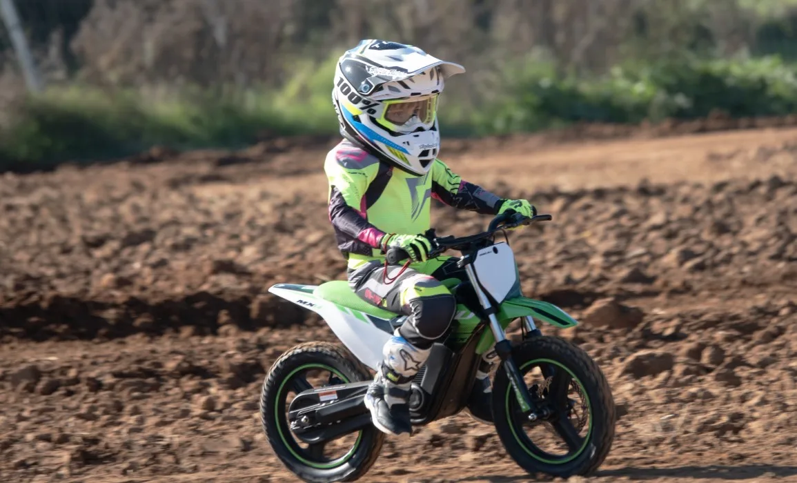 Kind auf MX14 500W E-Kindermotocross beim Fahren auf einer schlammigen Strecke mit Motocross-Ausrüstung.