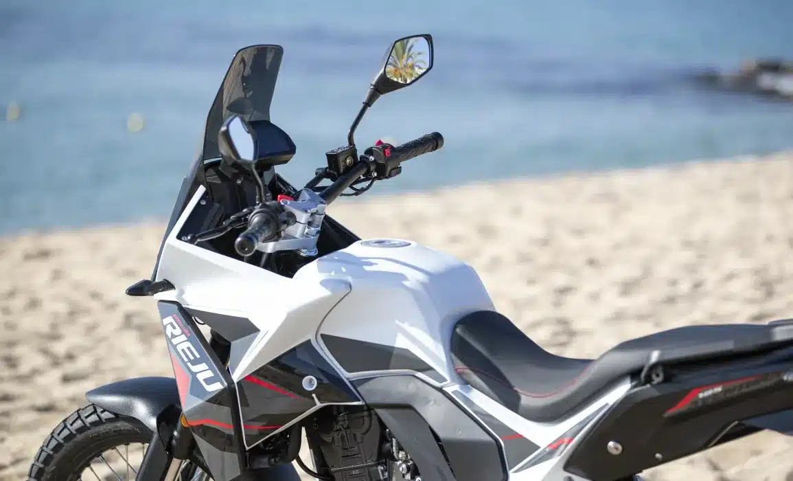 Rieju Aventura 125 Motorrad in Seitenansicht am Sandstrand, mit Blick auf den Lenker und das moderne Cockpit.