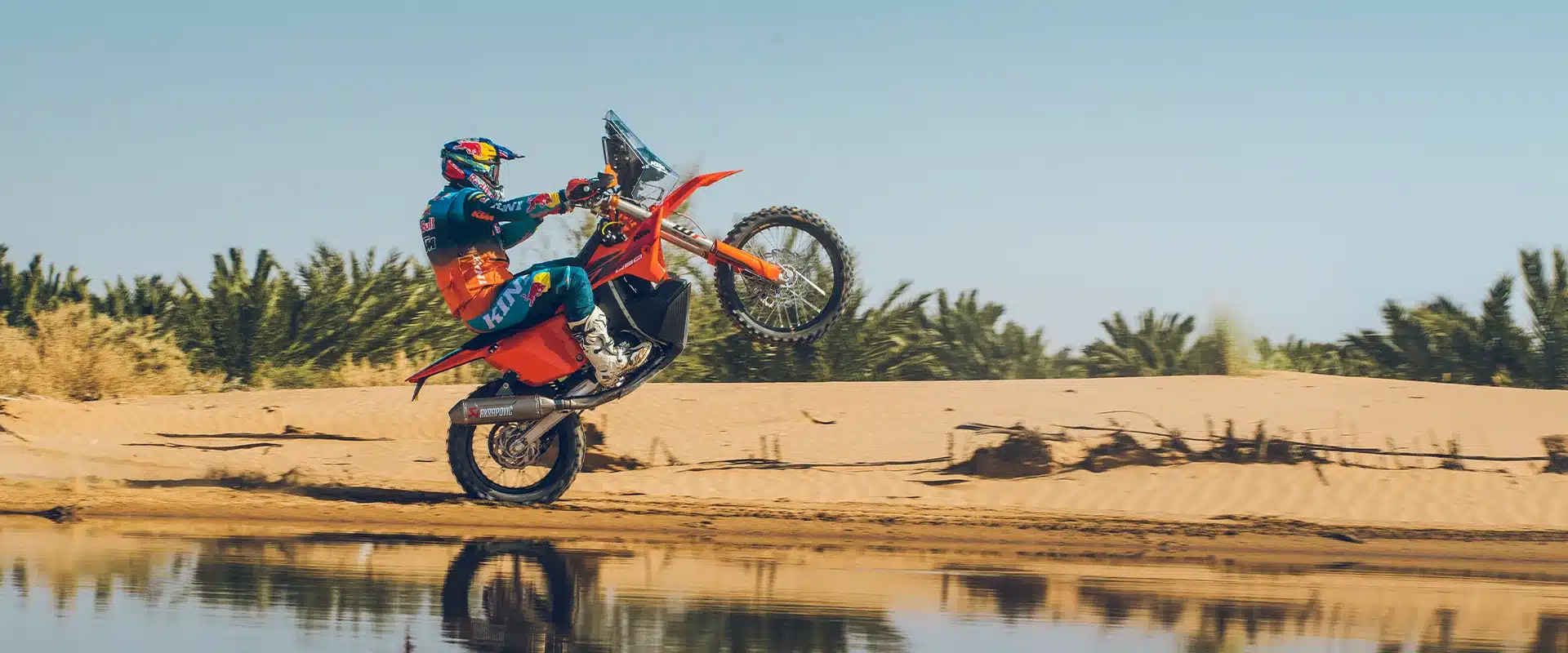 KTM 450 RALLY REPLICA 2027 beim Wheelie auf sandiger Strecke, Rallyefahrer in Ausrüstung.