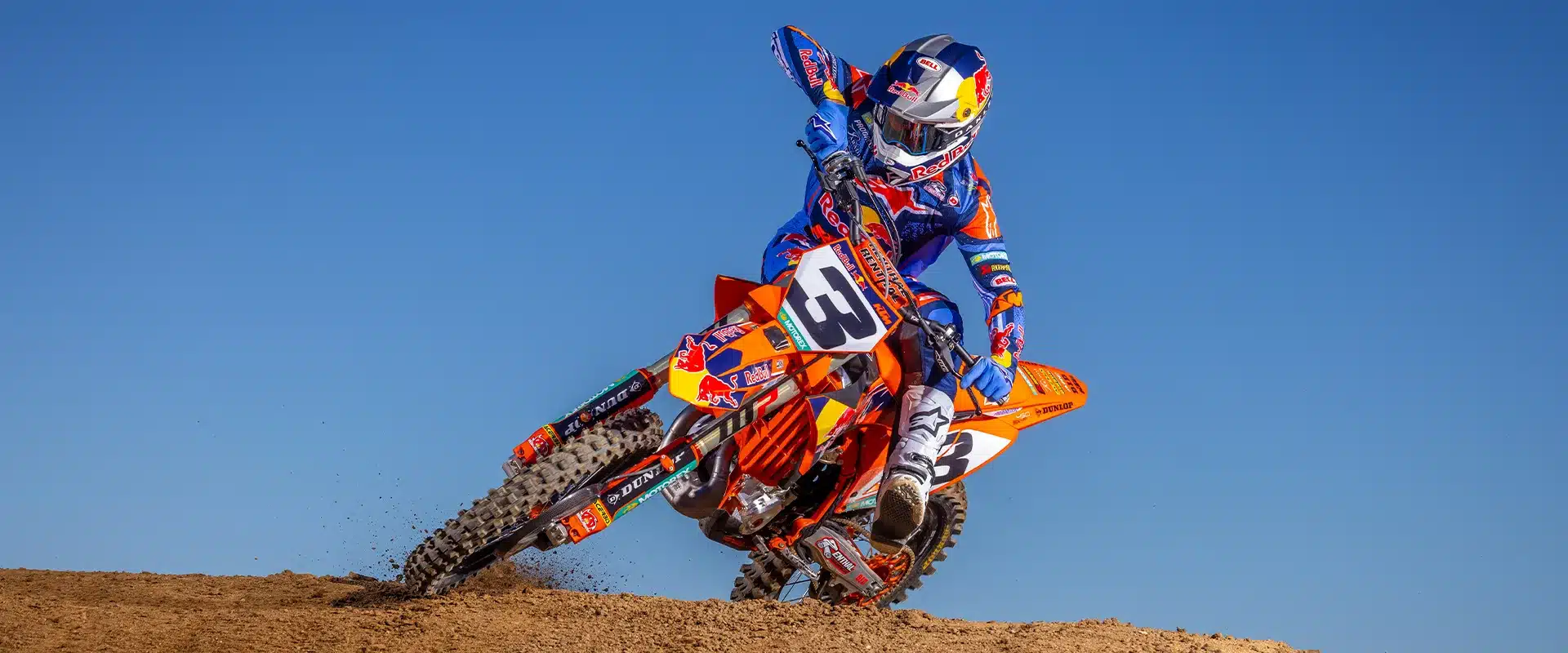 Motocross-Fahrer auf einer orangefarbenen KTM 450 SX-F Factory Edition 2026 mit Red-Bull-Grafik und Dunlop-Bereifung beim sportlichen Drift auf einer MX-Strecke.