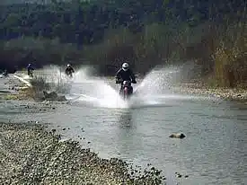 Mehrere Endurofahrer fahren durch einen Bach in den Bergen, Wasser spritzt hoch auf, vegetationsreiche Hänge im Hintergrund.