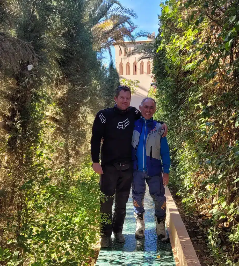 Zwei Männer in Enduro-Bekleidung stehen in einem mediterranen Garten zwischen Palmen und Pflanzen