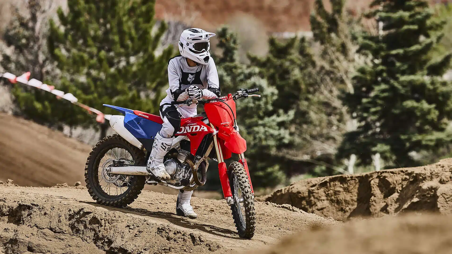 Fahrer sitzt auf einer Honda CRF450R Motocross-Maschine auf einer staubigen Rennstrecke mit Bäumen im Hintergrund.