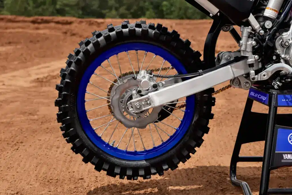 Nahaufnahme des Hinterrads mit blauer Felge und Kettenantrieb der Yamaha YZ85 (2027) auf sandigem Untergrund.