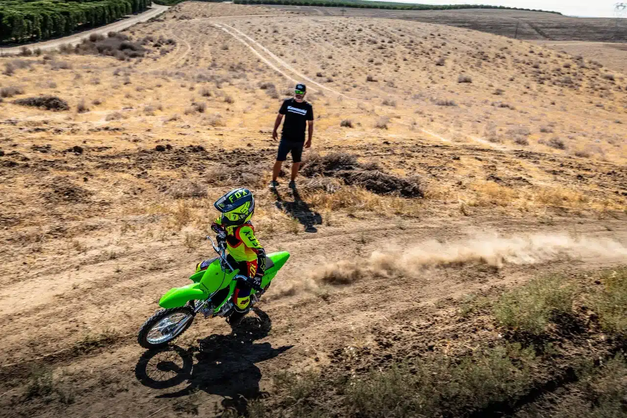 Kind auf grüner Kawasaki KLX110R fährt auf einer trockenen Offroad-Strecke, Betreuer im Hintergrund