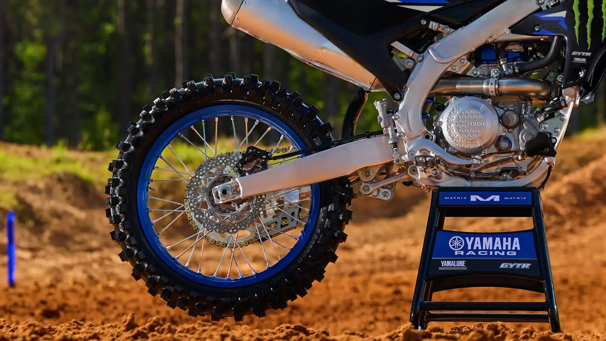 Detailansicht des Motors und Hinterrads der Yamaha YZ450F Monster Energy Yamaha Racing Edition, abgestellt auf einem Ständer auf sandigem Untergrund.