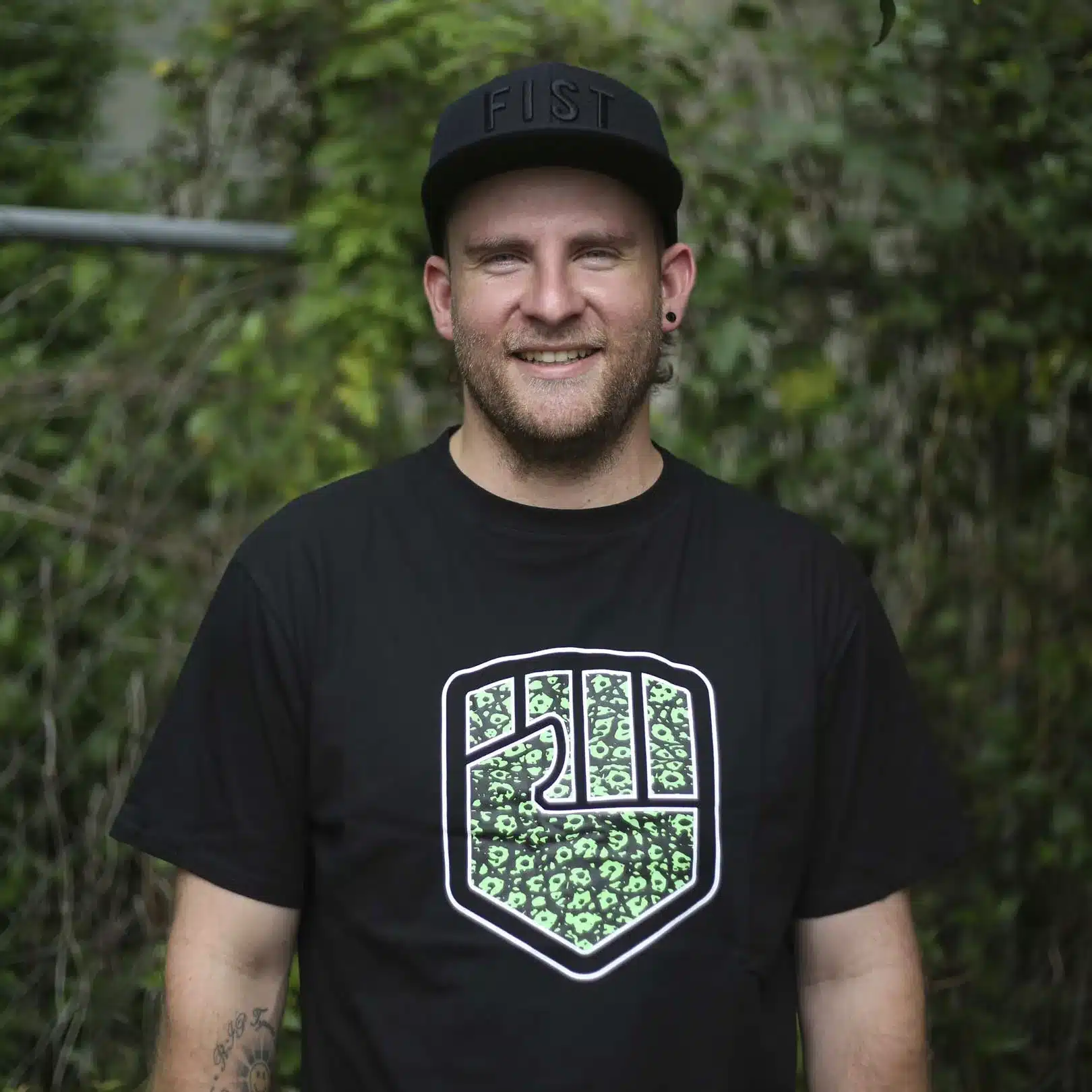 Mann trägt FIST Handwear CROC TEE, schwarzes T-Shirt mit grünem Logo, vor natürlichem Hintergrund.