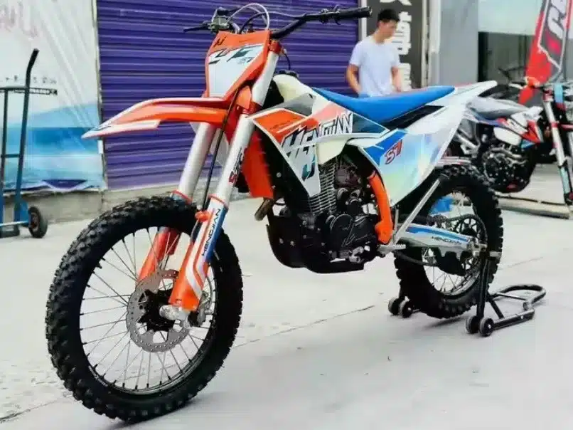 Hengjian S7-YBS300 Enduro-Motorrad in orange-weiß-blauer Lackierung auf einem Ständer vor einer Werkstatt.