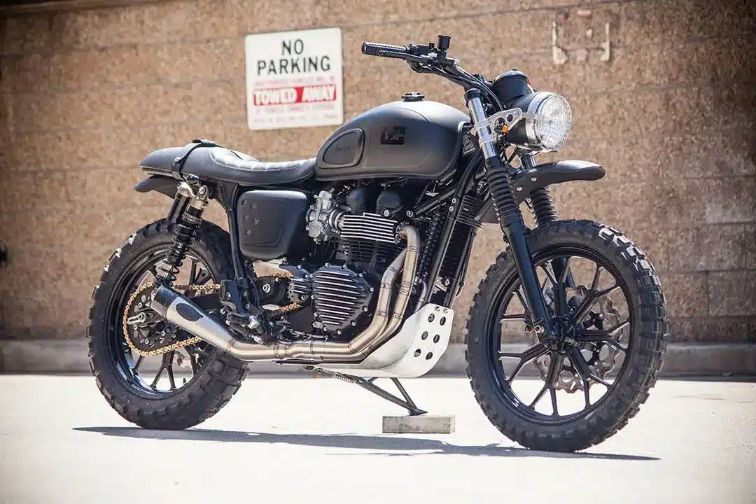 Schwarze Triumph Scrambler im Custom-Umbau mit groben Stollenreifen und schwarzem Aluminium-Finish, parkend vor einer Backsteinwand.