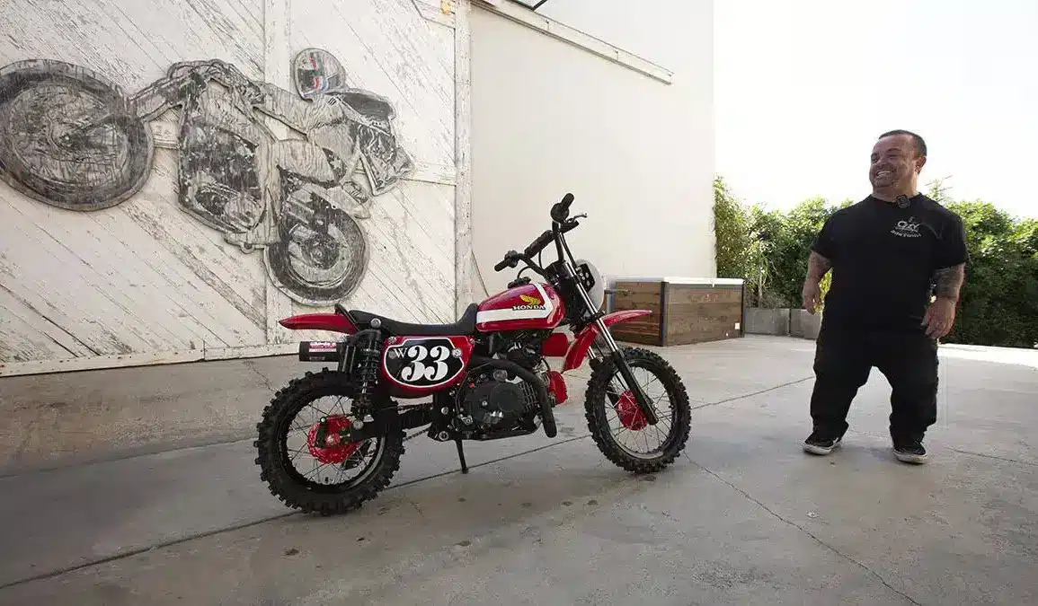 Wee Man CRF 110 Custom-Bike mit Fahrer im Hof vor Garagentor und Motorradkunstwerk im Hintergrund.