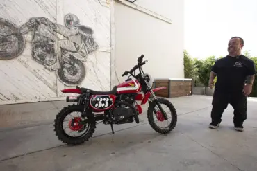 Wee Man CRF 110 Custom-Bike mit Fahrer im Hof vor Garagentor und Motorradkunstwerk im Hintergrund.