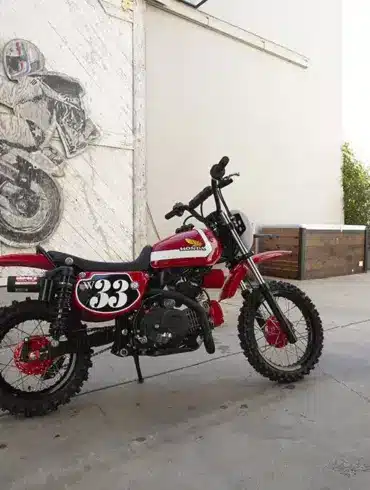 Wee Man CRF 110 Custom-Bike mit Fahrer im Hof vor Garagentor und Motorradkunstwerk im Hintergrund.