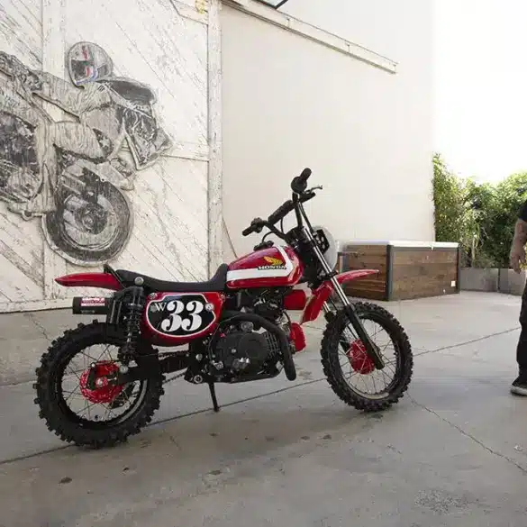 Wee Man CRF 110 Custom-Bike mit Fahrer im Hof vor Garagentor und Motorradkunstwerk im Hintergrund.