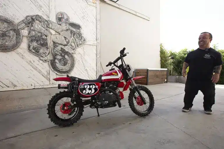 Wee Man CRF 110 Custom-Bike mit Fahrer im Hof vor Garagentor und Motorradkunstwerk im Hintergrund.