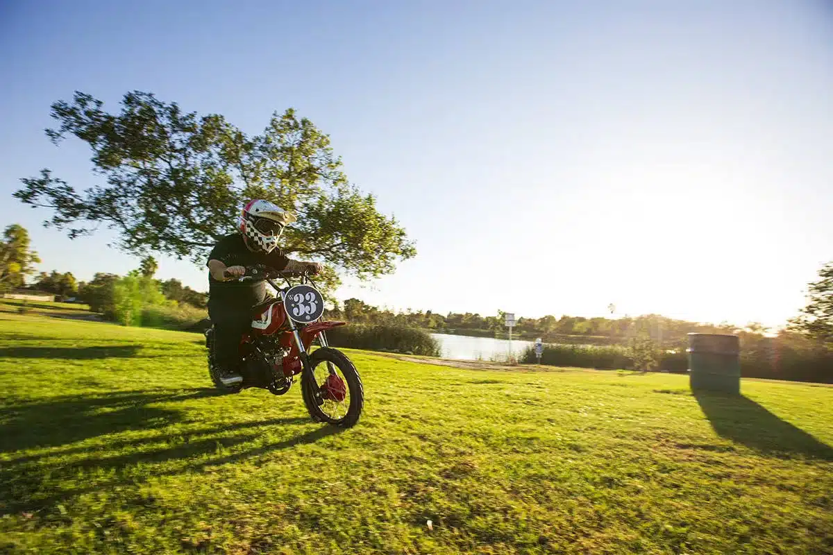 Fahrer mit Helm auf der umgebauten Wee Man CRF 110 im Retro-Stil fährt auf einer sonnigen Wiese.