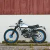 Seitenansicht einer individuell umgebauten Hodaka Wombat 'Blue Wombat' mit blauen Felgen, Aluminiumtank und Offroad-Bereifung vor einer roten Holzwand.