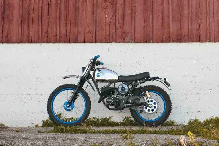 Seitenansicht einer individuell umgebauten Hodaka Wombat 'Blue Wombat' mit blauen Felgen, Aluminiumtank und Offroad-Bereifung vor einer roten Holzwand.