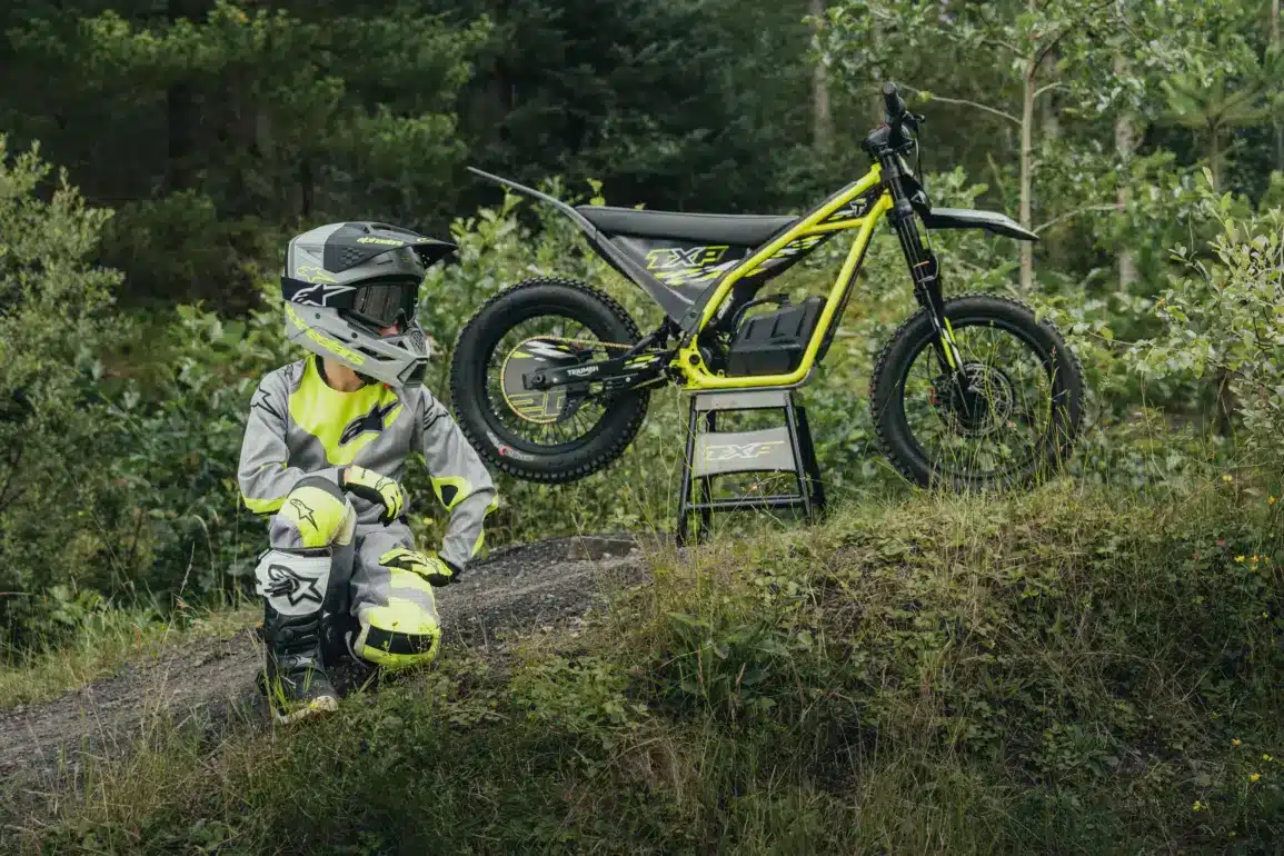 Ein Kind in Motocross-Schutzkleidung neben einer Triumph TXP-16 im Gelände.
