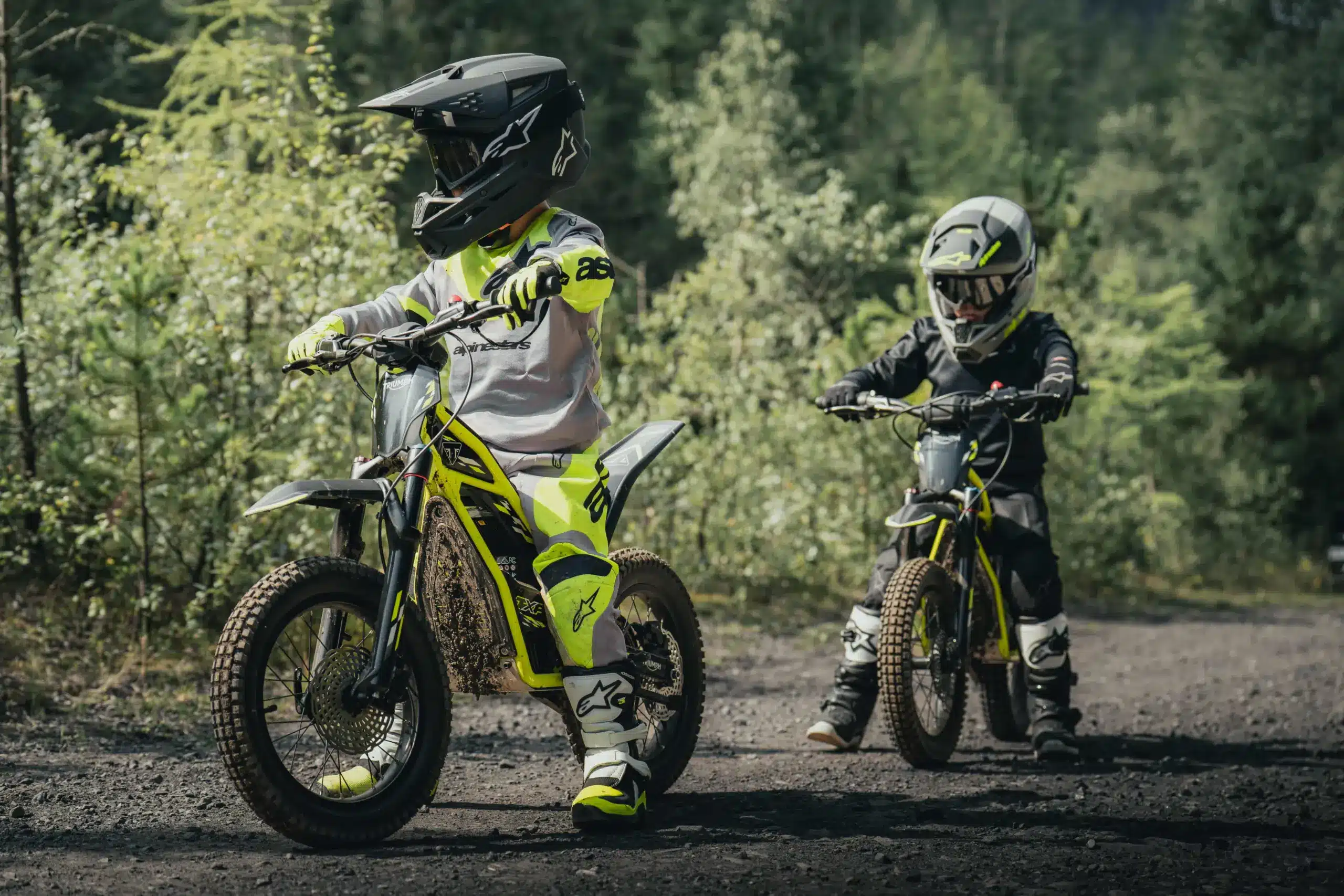 Zwei junge Fahrer auf Triumph TXP-16 Elektromotorrädern im Offroad-Gelände, ausgestattet mit Schutzkleidung und Helmen