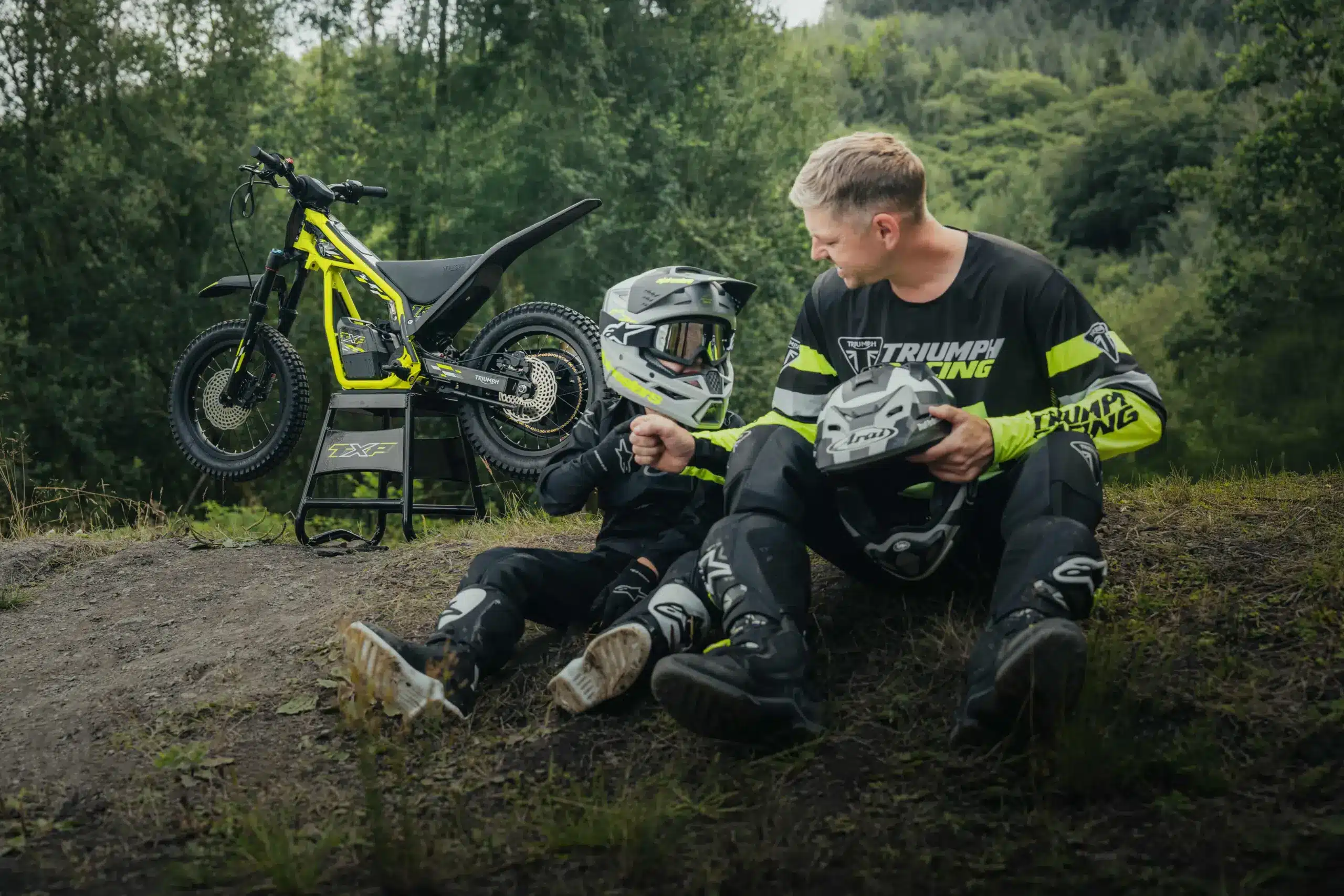 Ein Kind in Offroad-Ausrüstung mit erwachsenem Begleiter neben einer Triumph TXP-16, einem elektrischen Kinder-Offroadmotorrad, auf einer natürlichen Geländestrecke.
