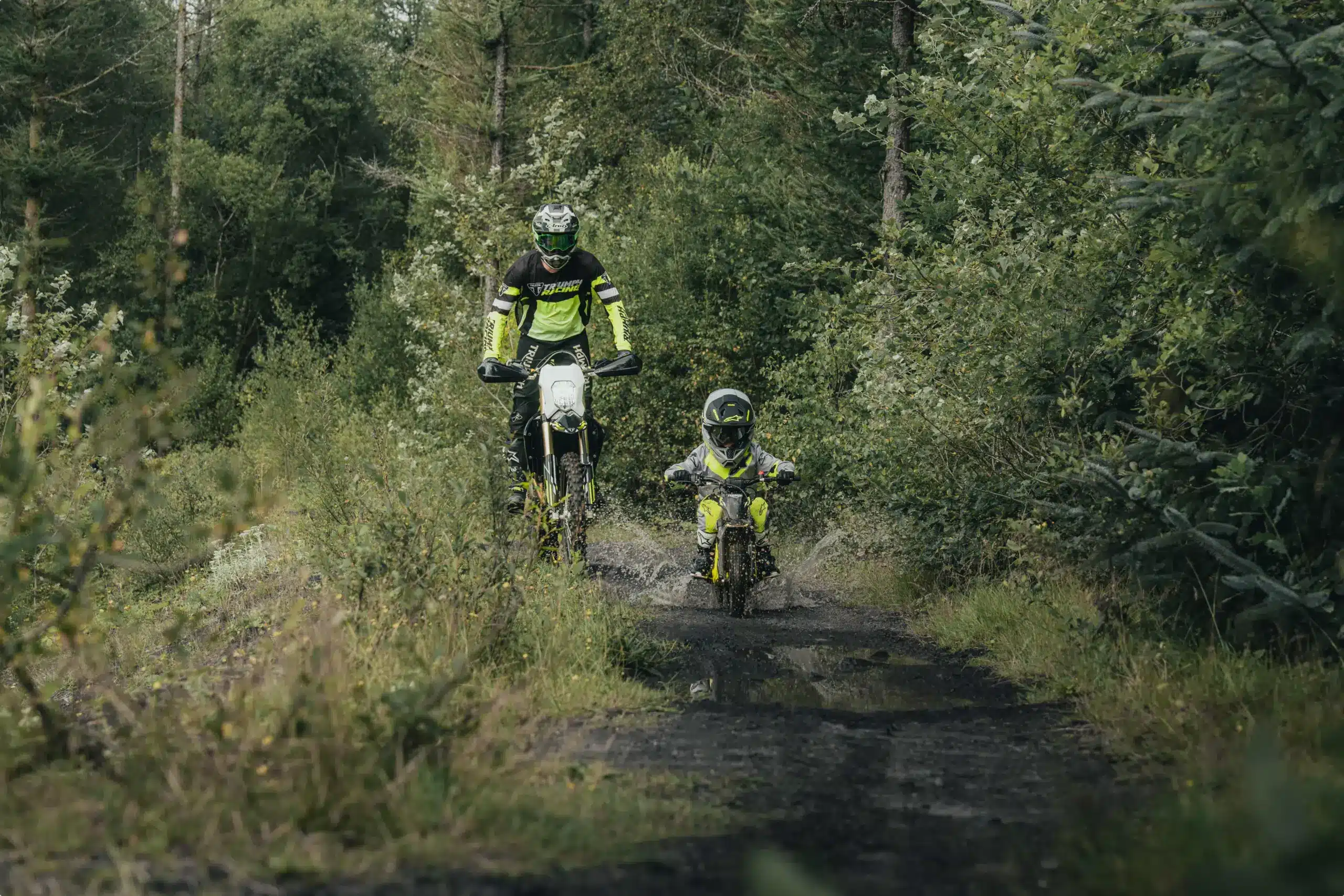 Kind auf Triumph TXP-12 Elektromotorrad zusammen mit Erwachsenem im Wald auf einem Offroad-Pfad.