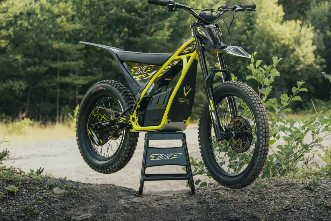 Triumph TXP-24 Elektromotorrad im Offroad-Einsatz auf einem Ständer vor Waldkulisse