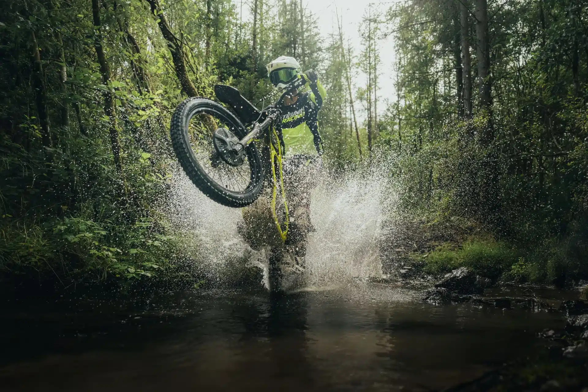 Triumph TXP-24 E-Offroad-Motorrad fährt dynamisch durch Wasser im Wald.